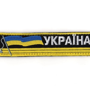 Puede incluir: Parche bordado con la bandera ucraniana en azul y amarillo, y la palabra "УКРАЇНА" en blanco. El parche tiene un fondo negro con un borde y costuras amarillas. La bandera ondea y el parche es rectangular.