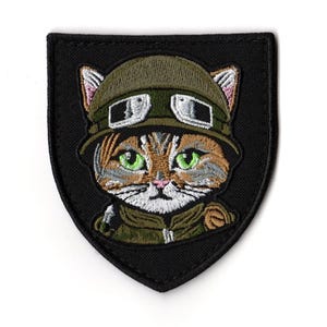 Puede incluir: Parche bordado con un gato con casco y gafas de estilo militar. El gato tiene ojos verdes y pelaje marrón y blanco. El parche es negro y tiene forma de escudo.