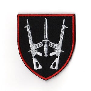 Puede incluir: Parche negro con borde rojo, con un diseño bordado en blanco. El diseño incluye dos rifles que flanquean una espada central. El parche tiene forma de escudo y es adecuado para ropa militar o táctica.