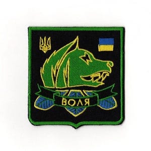 Puede incluir: Un parche cuadrado con fondo negro y borde verde. El parche presenta una cabeza de lobo verde con contornos amarillos, una pancarta con la palabra "VOLYA" y una pequeña bandera ucraniana. El parche también incluye un símbolo de tridente amarillo.