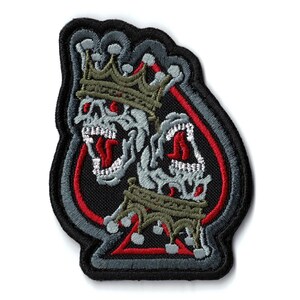 May include: Embroidered patch featuring two skull faces wearing crowns, set against a black spade shape. The skulls have red eyes and open mouths, with a red outline. The patch has a black, gray, and olive green color scheme.