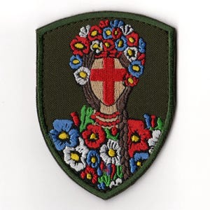 Puede incluir: Parche bordado que representa a una mujer con una cruz roja en la cara, adornada con una corona floral y un ramo. El parche tiene forma de escudo, verde con un borde verde. Las flores son rojas, azules y blancas.