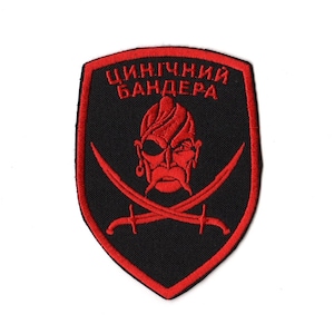Puede incluir: Parche bordado con forma de escudo negro y borde rojo. El parche presenta una cara roja de pirata con bigote y parche en el ojo, espadas cruzadas y texto cirílico.