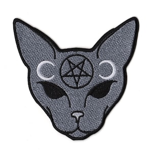 May include: A grey embroidered patch in the shape of a cat head with large pointed ears. The design features a black pentagram centered on the forehead, flanked by two white crescent moons. The cat has solid black almond-shaped eyes and a black border.