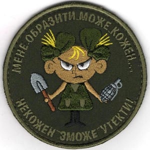 Puede incluir: Parche bordado circular con una niña de dibujos animados enfadada, sosteniendo una pala y una granada. El parche es principalmente verde oliva, marrón y amarillo, con texto en idioma extranjero alrededor del borde.