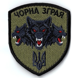 Puede incluir: Parche bordado con forma de escudo, con tres cabezas de lobo negras con ojos rojos y bocas abiertas. El texto "ЧОРНА ЗГРАЯ" está arqueado sobre los lobos. El parche tiene un fondo verde y un borde negro.