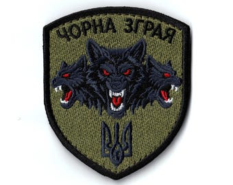 May include: Embroidered patch with a shield shape, featuring three black wolf heads with red eyes and open mouths. The text "ЧОРНА ЗГРАЯ" is arched above the wolves. The patch has a green background and a black border.