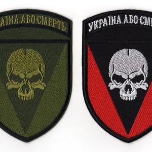 Puede incluir: Dos parches bordados con forma de escudo. Uno es verde oliva con una calavera y el texto "UKRAINA ABO SMERT" en blanco. El otro es negro con un cheurón rojo, una calavera blanca y el mismo texto.