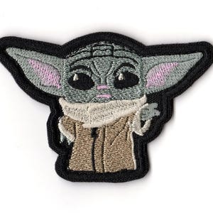 May include: Embroidered patch featuring a stylized depiction of a Grogu character from the Star Wars franchise. The patch has a black border, with the character in shades of gray, tan, and pink. The character is in a pose with one hand raised.