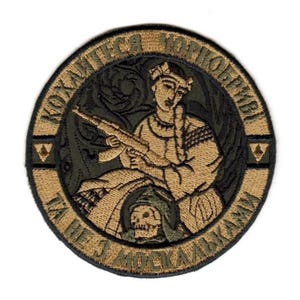 Puede incluir: Parche bordado redondo con un diseño central que presenta una figura sosteniendo un rifle, rodeada de texto. La paleta de colores incluye tonos de verde, beige y negro. El parche tiene una apariencia detallada y texturizada.