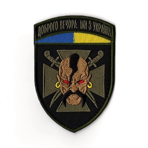 Puede incluir: Parche bordado con un diseño de escudo, que incorpora la bandera ucraniana en la parte superior. Presenta la cara estilizada de un guerrero con bigote, pendientes y espadas cruzadas. El texto "Доброго вечора. Ми з України" está arqueado arriba.