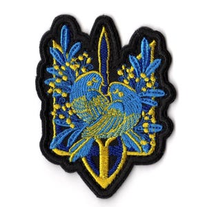 Könnte beinhalten: Gestickter Aufnäher mit einem blau-gelben Vogel- und Blumendesign auf schwarzem Hintergrund. Der Aufnäher hat einen gewellten Rand. Geeignet für Kleidung oder Accessoires.