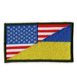 Puede incluir: Parche bordado que combina las banderas estadounidense y ucraniana. El parche rectangular tiene un borde verde. La sección de la bandera estadounidense muestra estrellas y rayas, mientras que la bandera ucraniana está representada por azul y amarillo.