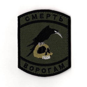 Puede incluir: Parche bordado con un cuervo negro posado sobre una calavera. El parche tiene un fondo verde con bordes negros y texto en una fuente estilizada. El texto dice "СМЕРТЬ ВОРОГАМ".