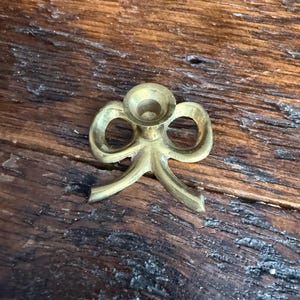 May include: A polished brass candle holder shaped like a decorative bow. The object features a central circular cup for holding a taper candle, flanked by two symmetrical loops and two downward-curving tails, all crafted from a single piece of metal.