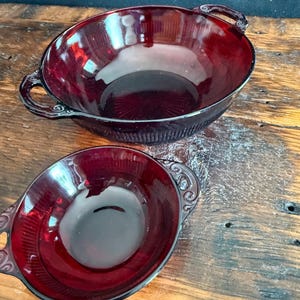 May include: Two deep red glass bowls with handles on each side. The bowls feature a vertical ribbed pattern along the exterior walls and a smooth interior surface. The handles are decorated with a swirling scroll design on the outer edges.