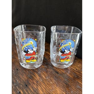 May include: Two clear square-shaped glasses with a Mickey Mouse graphic. The graphic features Mickey Mouse in a blue hat, red shorts, and yellow shoes, with a fireworks design. The number "2000" is printed below the graphic.