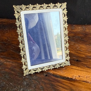 May include: A rectangular picture frame featuring an ornate, metallic gold-toned border with a repeating fleur-de-lis pattern. The inner edge of the frame is finished with a thin, silver-toned beaded trim surrounding the clear glass pane.