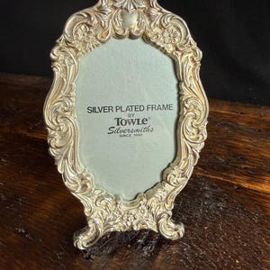 May include: An oval silver-plated picture frame with ornate floral and scrollwork relief detailing. The center insert features the text: SILVER PLATED FRAME BY TOWLE Silversmiths SINCE 1690. The frame has a pointed top and stands on two feet.