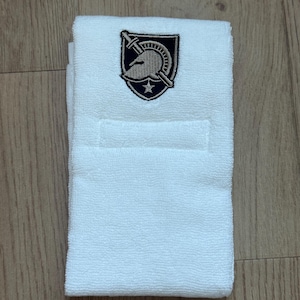 Puede incluir: Una toalla de mano blanca doblada con un emblema bordado. El emblema presenta un casco, una espada y una estrella dentro de un escudo en negro, dorado y azul marino. La toalla está hecha de un material suave y absorbente.