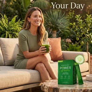 A beautiful outdoor lifestyle photo titled "Nourish Your Day." A smiling woman is sitting on a sofa in a sunlit garden, holding a glass of green juice. A box of Power Greens is on a wooden table next to a fresh cucumber slice, highlighting a natural and healthy daily routine.