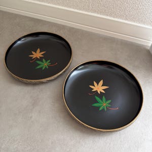 以下が含まれることがあります： 黒い丸いお盆が2つ。金色の縁取りがあり、それぞれに金と緑の葉と赤いアクセントのデザインが施されています。お盆は明るい灰色の表面に置かれています。