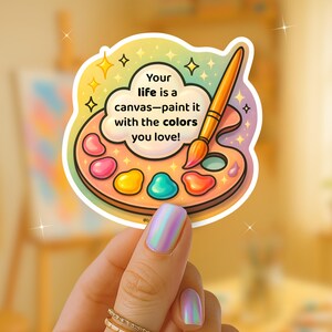 May include: A colorful sticker featuring a painter's palette with various paint colors and a paintbrush. The sticker has the text "Your life is a canvas-paint it with the colors you love!" in a cloud-shaped speech bubble. The sticker is held by a hand with iridescent nails.