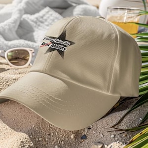 Peut inclure: Une casquette de baseball beige avec un motif d'étoile noire sur le panneau avant. À l'intérieur de l'étoile, le texte "Please Notice My Dis Ability" est écrit dans une police stylisée, avec une fine ligne rouge barrant le mot "Dis".