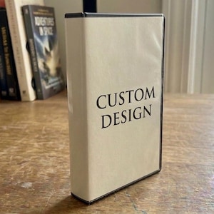 May include: A rectangular, off-white case with the words "CUSTOM DESIGN" printed in black. The case has a black border and is standing upright on a wooden surface. Books are visible in the background.