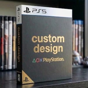 Könnte beinhalten: Eine schwarze PS5-Spielhülle mit goldfarbenem Schriftzug "custom design" und dem PlayStation-Logo. Die Hülle hat eine weiße Oberseite mit dem PS5-Logo. Auf dem Buchrücken steht vertikal "custom design". Weitere Spielhüllen sind im Hintergrund.