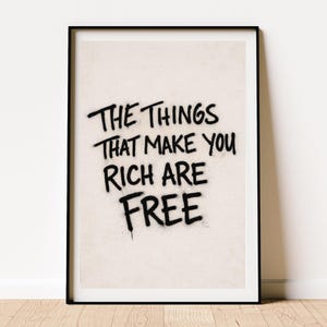 Könnte beinhalten: Gerahmter Kunstdruck mit dem Text "THE THINGS THAT MAKE YOU RICH ARE FREE" in schwarzer Graffiti-Schrift. Der Text befindet sich auf einem hellbeigen Hintergrund, der von einem schwarzen Rahmen umschlossen ist.