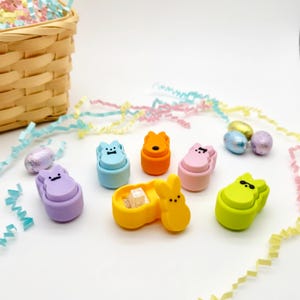 Puede incluir: Un surtido de coloridas gomas de borrar con temática de Pascua en forma de conejitos y pollitos. Las gomas de borrar son de varios colores, incluyendo morado, azul, naranja, rosa y verde. Un recipiente amarillo contiene pequeñas gomas blancas. Una cesta de Pascua decorativa y serpentinas de papel de colores están en el fondo.