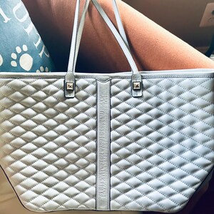 May include: A light gray tote bag featuring a quilted diamond pattern across the front panels. The bag includes a smooth vertical center strip, two thin shoulder straps, and two small square metallic studs at the base of the handle attachments.