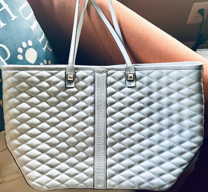 May include: A light gray tote bag featuring a quilted diamond pattern across the front panels. The bag includes a smooth vertical center strip, two thin shoulder straps, and two small square metallic studs at the base of the handle attachments.