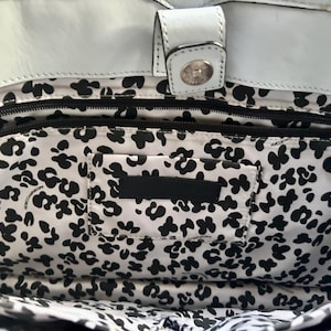 May include: The interior of a white leather bag featuring a black and white floral pattern lining. The bag includes a top magnetic snap closure, a horizontal zippered pocket, and a rectangular patch pocket centered on the patterned fabric interior.