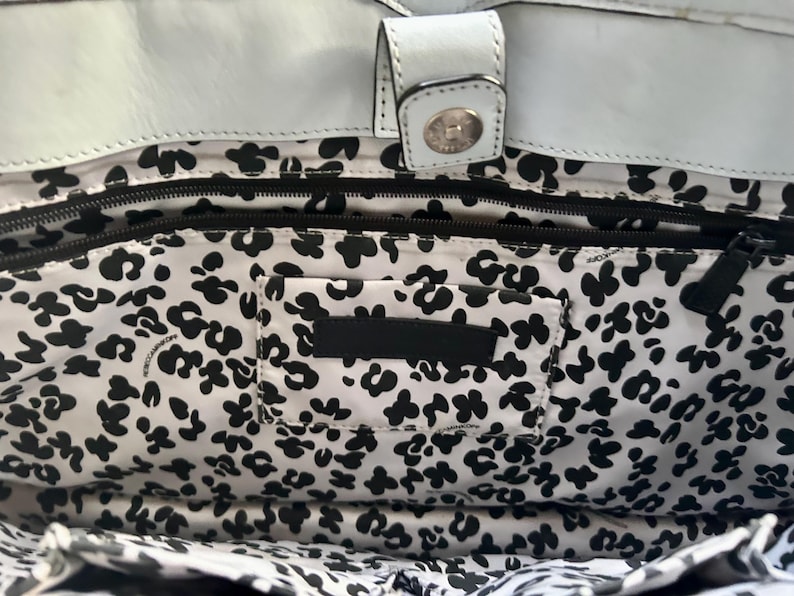 May include: The interior of a white leather bag featuring a black and white floral pattern lining. The bag includes a top magnetic snap closure, a horizontal zippered pocket, and a rectangular patch pocket centered on the patterned fabric interior.