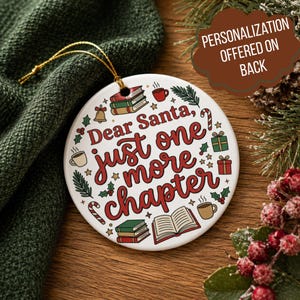 Może przedstawiać: Okrągła biała ceramiczna ozdoba z czerwonym napisem Dear Santa, just one more chapter. Wzór zawiera ilustracje stosów książek, otwartej książki, kubków do kawy, lasek cukrowych, dzwonków i liści ostrokrzewu ze złotym sznurkiem do zawieszenia.