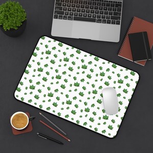 Pode incluir: Tapete de rato branco com borda preta, coberto por um padrão de folhas verdes. Um rato de computador branco está sobre o tapete. O tapete está sobre uma superfície escura, com um laptop, uma chávena de café e artigos de papelaria por perto.