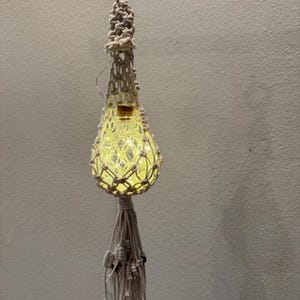 May include: A handmade macrame hanging light fixture. The light is encased in a woven, off-white macrame holder with a wooden ring at the top for hanging. The light inside glows with a warm, yellow hue, and the bottom has a decorative tassel.