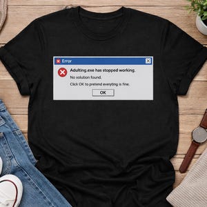 May include: Black t-shirt featuring a computer error message: "Adulting.exe has stopped working. No solution found. Click OK to pretend everything is fine." The shirt is displayed on a wooden surface with jeans, shoes, and accessories.