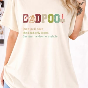 May include: Cream-colored t-shirt with the word "DADPOOL" in colorful block letters. The design includes a definition and the phrase "like a dad, only cooler." The shirt is a casual wear item.