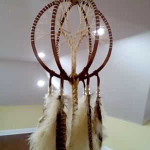 May include: A handcrafted dreamcatcher with a brown frame and a central tree-like design. The dreamcatcher features multiple circular hoops, adorned with feathers in shades of white and brown, and is suspended from a single point.