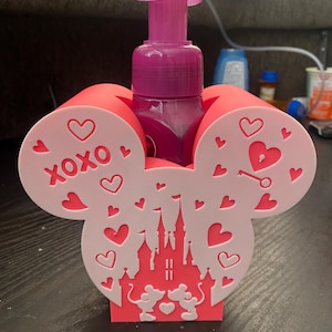 May include: A pink and white Mickey Mouse-shaped soap dispenser holder with a pink soap dispenser. The holder features hearts, the text "XOXO", and a castle design with Mickey and Minnie Mouse. The dispenser is a Valentine's Day theme.