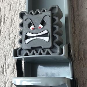 May include: A gray, 3D-printed Thwomp character from the Super Mario video game series. The Thwomp has an angry expression with red eyes and white teeth. The character is mounted on a gray metal object.