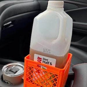May include: A clear plastic gallon jug of Diet Half & Half beverage sits in an orange plastic crate inside a vehicle. The jug has a white cap and a handle. The crate is in a car's cup holder area.