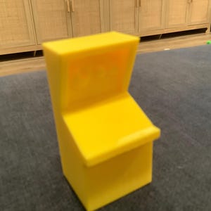 May include: A bright yellow plastic miniature arcade cabinet replica featuring a slanted control panel area and a recessed screen section. The object has a solid, blocky structure with smooth surfaces and a distinct upright design.