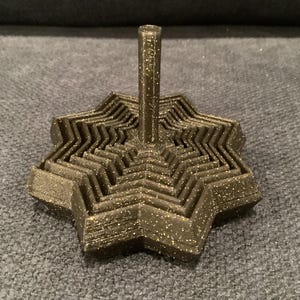 May include: A metallic gold-colored, star-shaped decorative object with a central vertical post. The object has a layered design with a zig-zag pattern around the star's perimeter. The surface has a textured appearance with small, glitter-like particles.