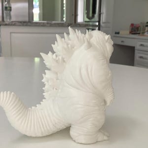 May include: A white, unpainted figurine of a monster with a textured, reptilian-like body, featuring a series of sharp, jagged dorsal fins running along its back and a thick, curved tail extending outward from its lower torso.