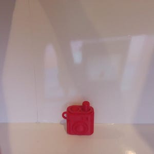 May include: A bright red silicone protective case for wireless earbuds, designed to resemble a vintage camera. The case features a circular lens-like detail on the front, a small loop on the side, and a top opening for the earbud charging port.