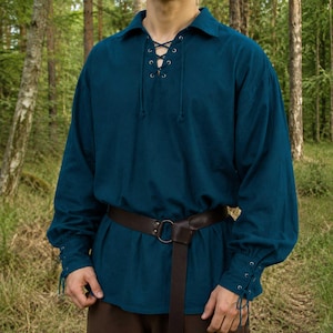 May include: A teal long-sleeved shirt featuring a lace-up front with metal eyelets and matching cord ties. The garment has a fold-down collar, gathered cuffs with lace-up details, and is worn with a dark brown leather belt featuring a metal ring.